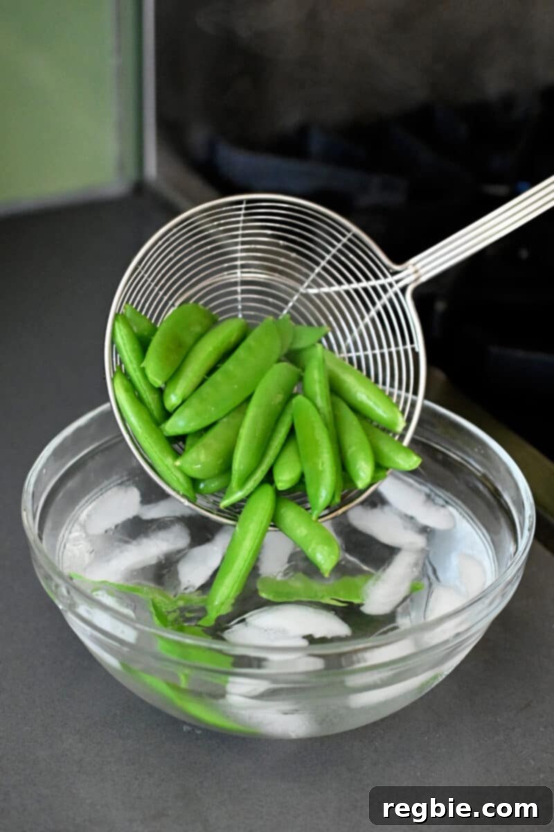 Whole30 Vegan Asian Chopped Salad 9 A metal strainer carefully lowering a batch of bright green, cooked sugar snap peas into a bowl brimming with ice water, to quickly chill them.