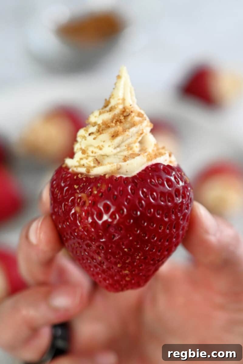 A hand is holding up a paleo and vegan cheesecake stuffed strawberry topped with gluten-free cookie crumbs, showcasing its creamy filling and vibrant color.