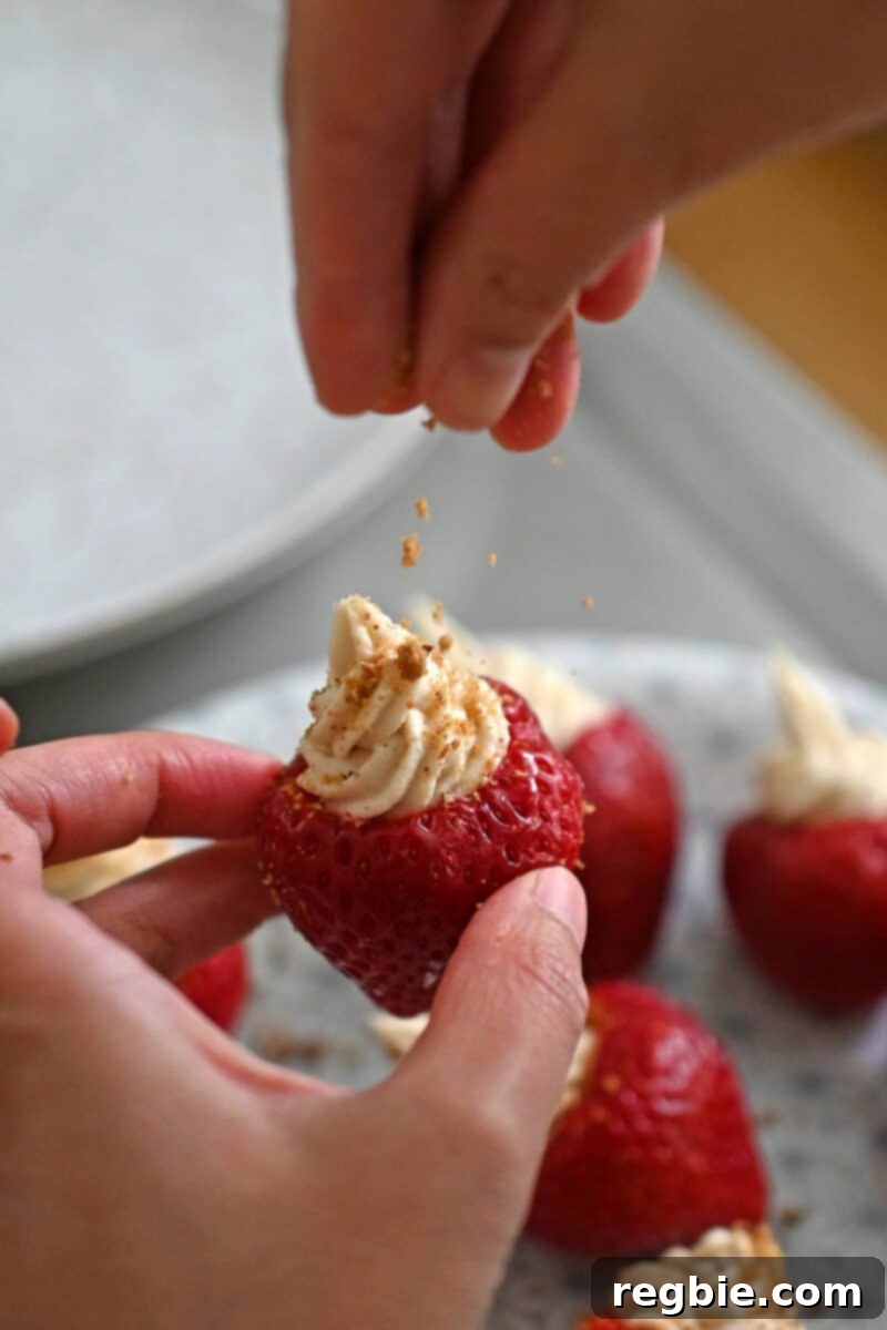 A hand is gently sprinkling crushed paleo cookie crumbs over the top of a vegan and dairy-free cheesecake stuffed strawberry, creating an appetizing finish.