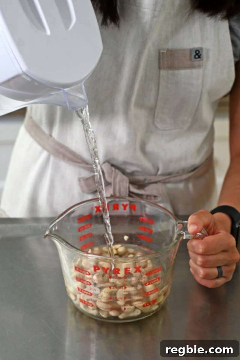 A close-up shot of water being poured into a clear glass measuring cup filled with raw cashews, initiating the soaking process.