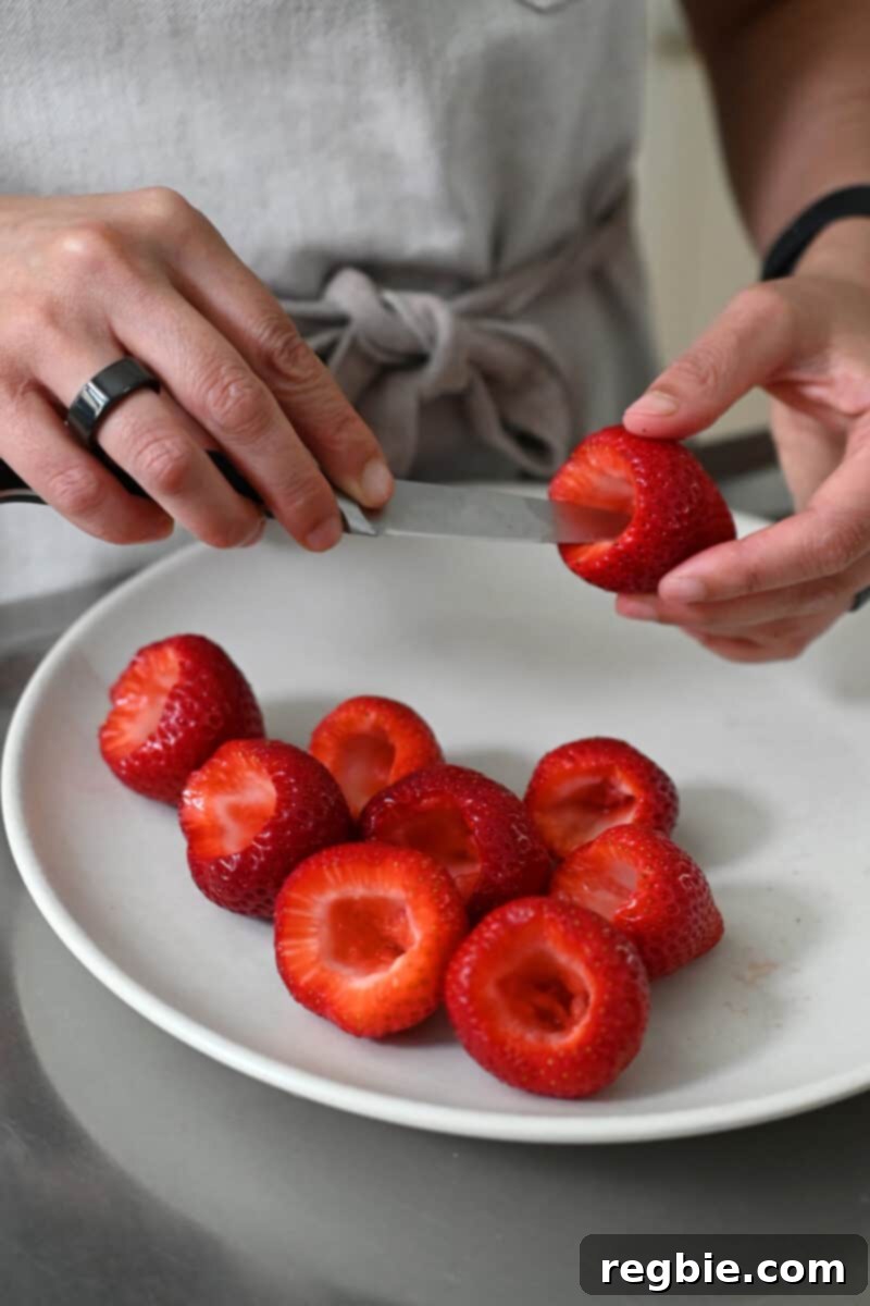 A small paring knife is precisely hollowing out a fresh strawberry on a plate that is already filled with several other hollowed-out strawberries, demonstrating the preparation step.