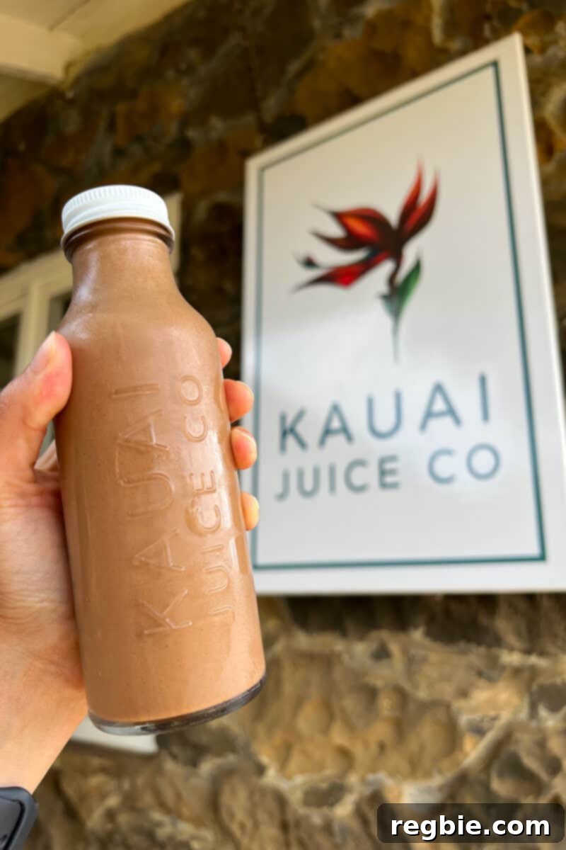 A hand gracefully holds a bottle of Black Pearl drink from Kauai Juice Co in front of a rustic store sign, illustrating their fresh beverage offerings.