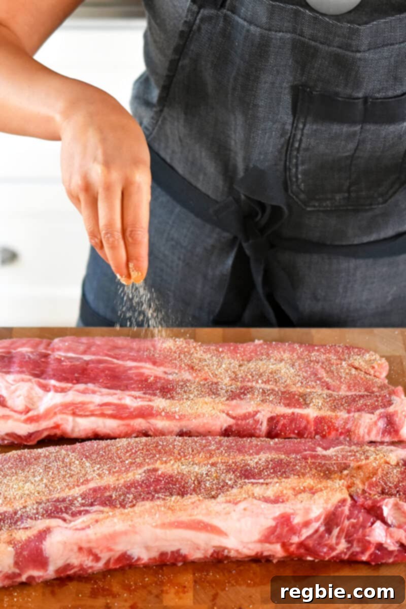 A person in a gray apron is sprinkling sugar-free dry rub on baby back ribs.