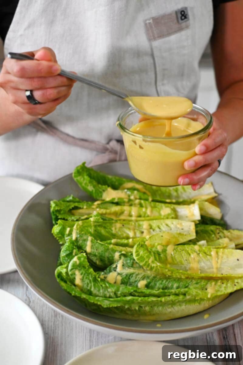 Creamy Dairy Free Caesar Dressing Whole30 Keto 3 Someone adding generous dollops of homemade dairy-free Caesar dressing to a shallow bowl filled with fresh romaine lettuce leaves, creating a vibrant salad.