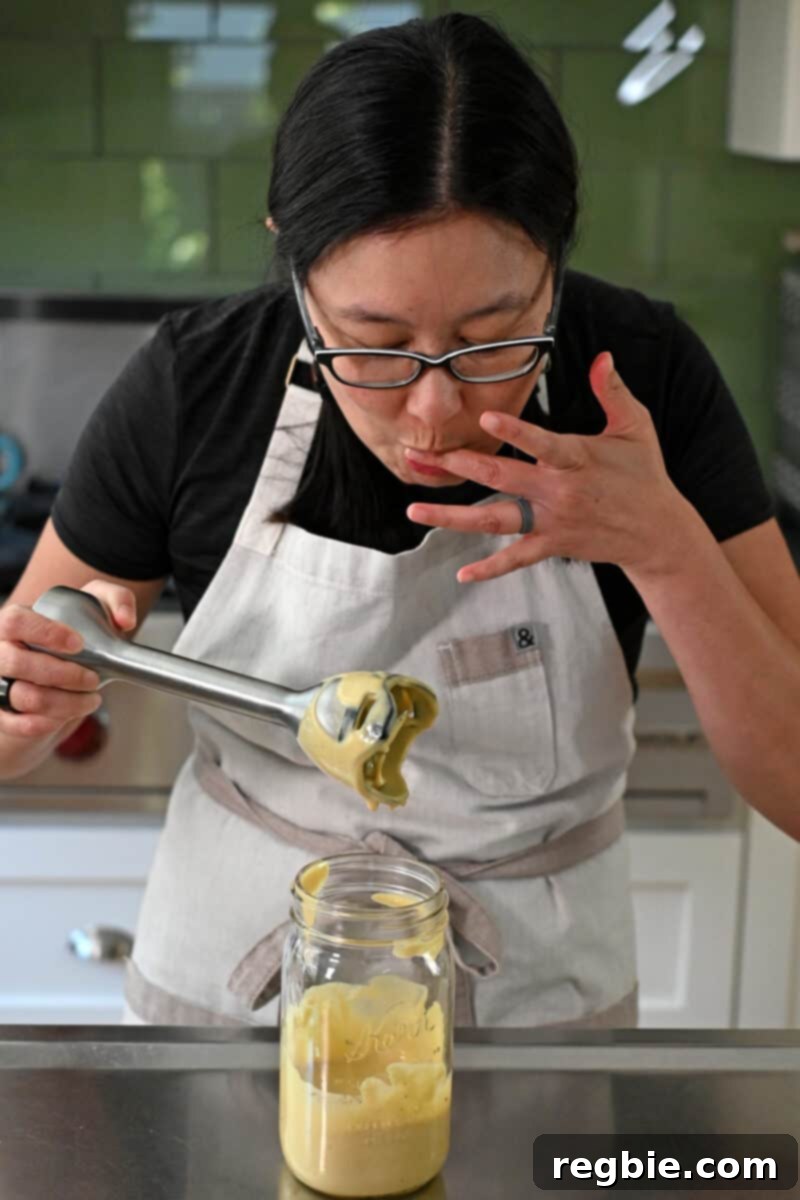 Creamy Dairy Free Caesar Dressing Whole30 Keto 7 An Asian woman with glasses is tasting a freshly blended batch of dairy-free Caesar dressing from a spoon, evaluating its flavor.