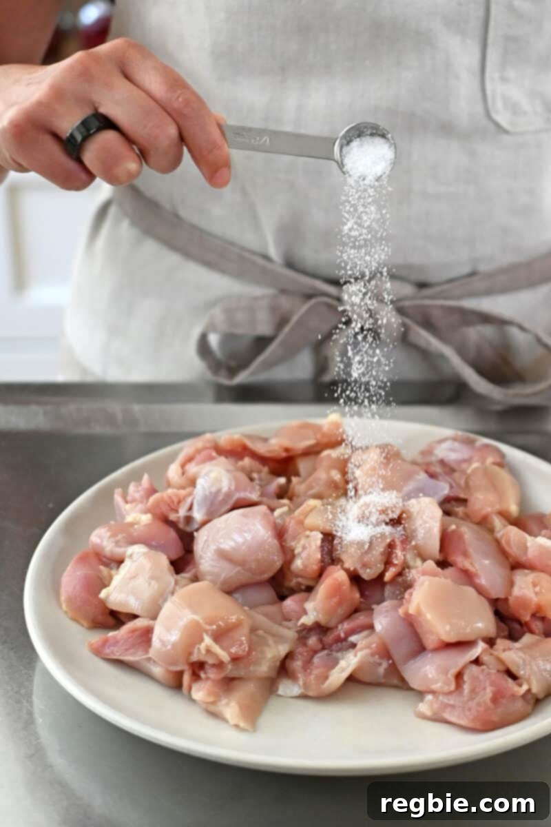 Healthy Instant Pot Chicken Saag Whole30 Keto 10 A person in an off-white apron is adding a small spoonful of salt to a plate filled with raw cut-up boneless, skinless chicken thighs.