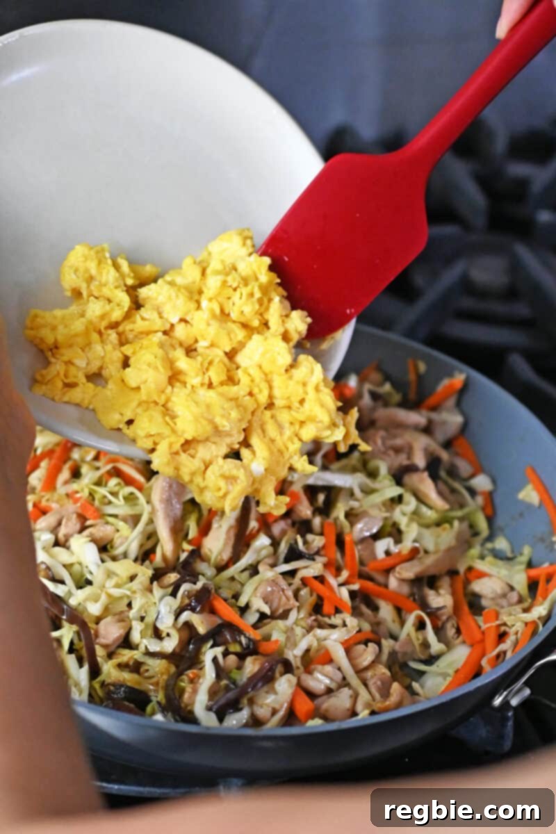 Adding a plate of cooked scrambled eggs into a skillet filled with Moo Shu Chicken, gently folding them in for the final touch.