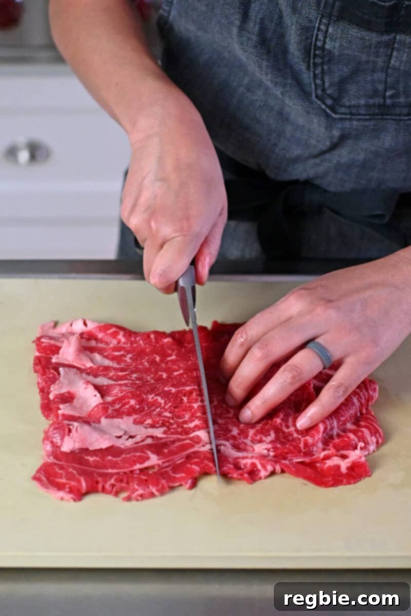 A person in an gray apron is slicing a stack of paper thin raw beef in half, crosswise for healthy gyudon.