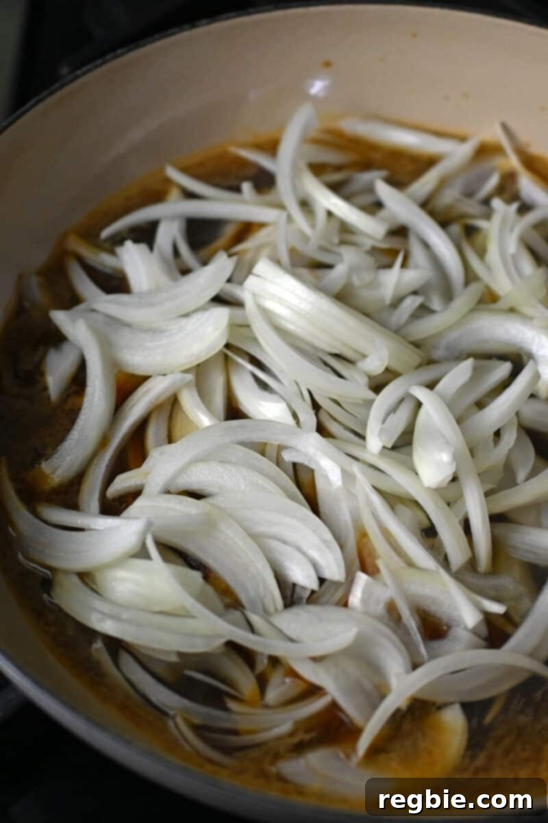 A closeup shot of raw thinly sliced onions in an enameled cast iron skillet in a brown sauce for healthy gyudon.