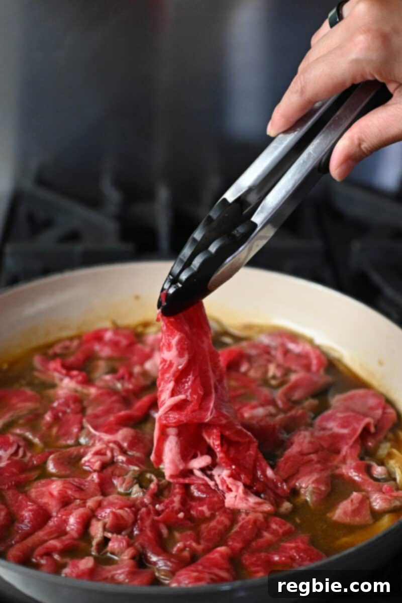 A pair of tongs is adding thinly sliced raw beef to a skillet filled with simmering brown sauce and sliced onions for healthy gyudon.