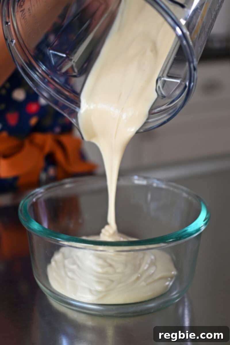 Pouring freshly blended vegan cream cheese frosting from a blender into a glass storage container for chilling.