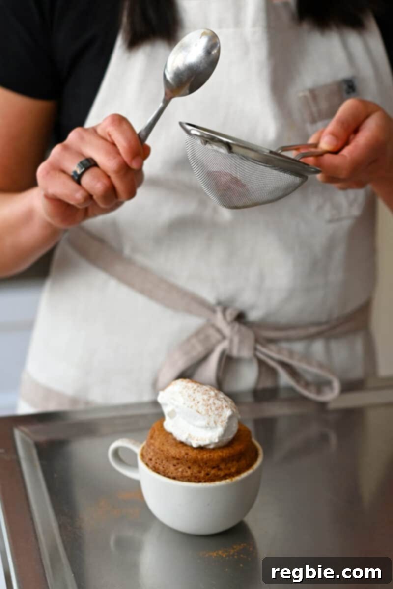 Gently tapping ground cinnamon through a fine mesh sieve onto an apple mug cake already topped with creamy coconut whipped cream, creating a beautiful finish.
