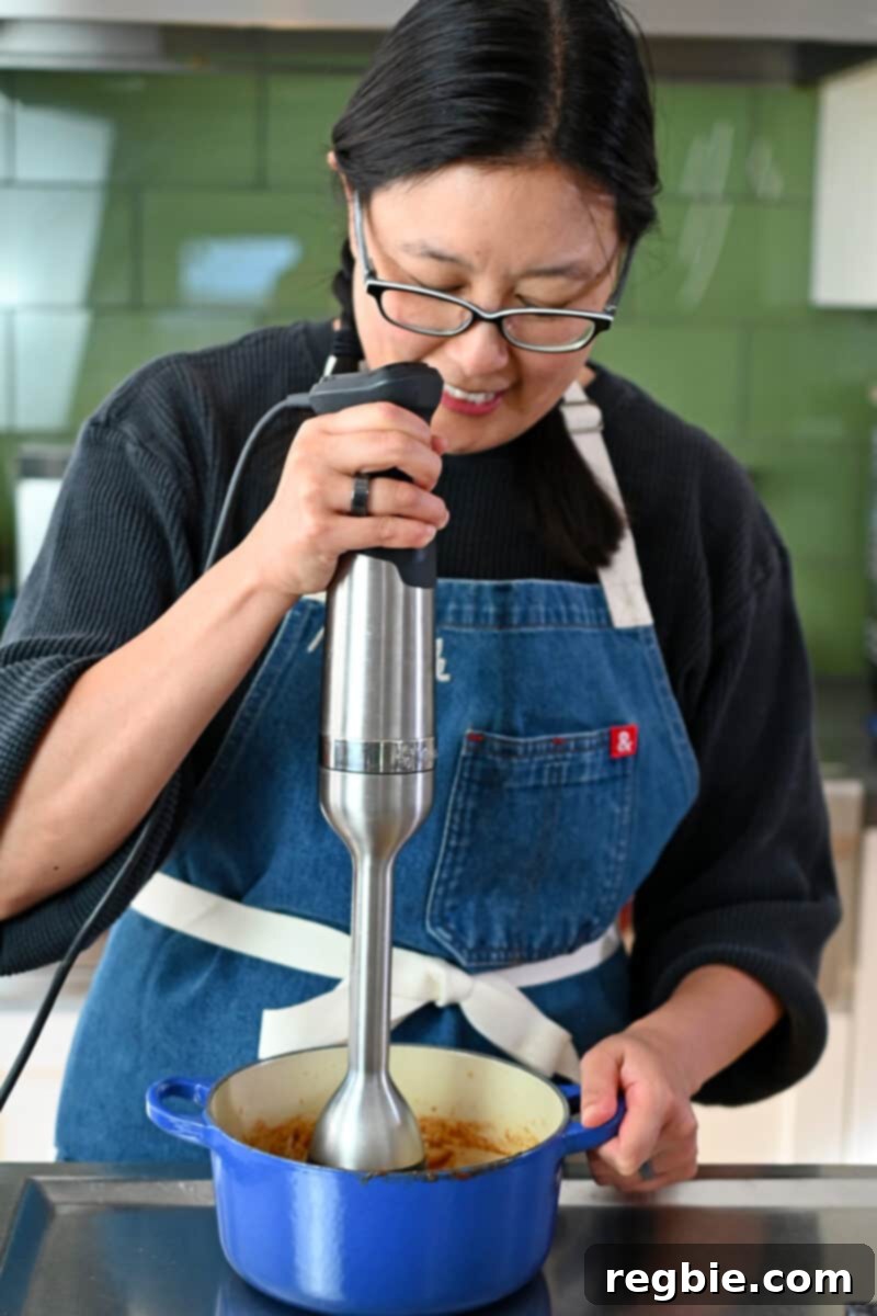 Healthy & Delicious Tonkatsu Sauce (Paleo, Whole30, Vegan) 11 A smiling Asian woman using an immersion blender to puree homemade Tonkatsu Sauce in a small blue saucepan, creating a smooth consistency.