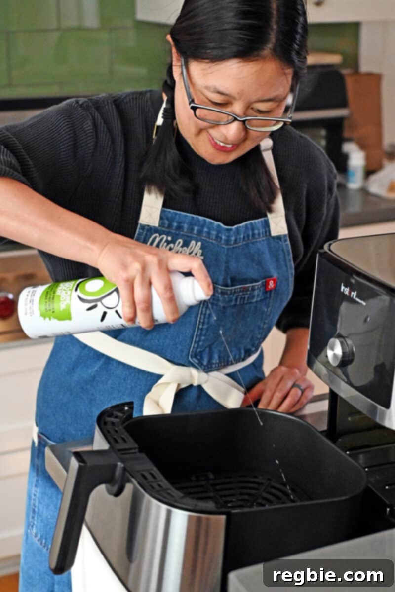 An Asian woman is spraying an open air fryer basket with avocado oil spray before placing the tonkatsu inside.