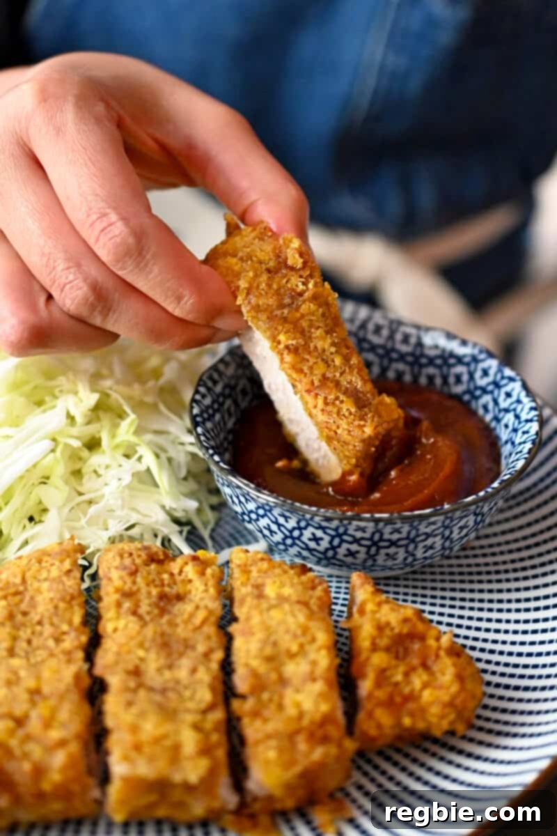 A hand dipping a perfectly golden slice of pork katsu into a small bowl filled with savory Tonkatsu sauce, highlighting the crispy crust.