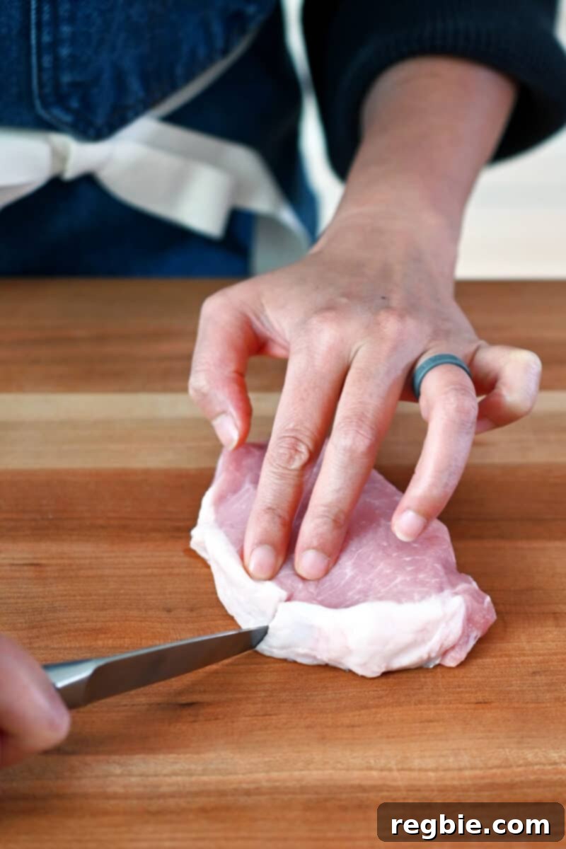 A paring knife is cutting a small slit in the connective tissue on the fatty side of a boneless pork chop to prevent curling during cooking.