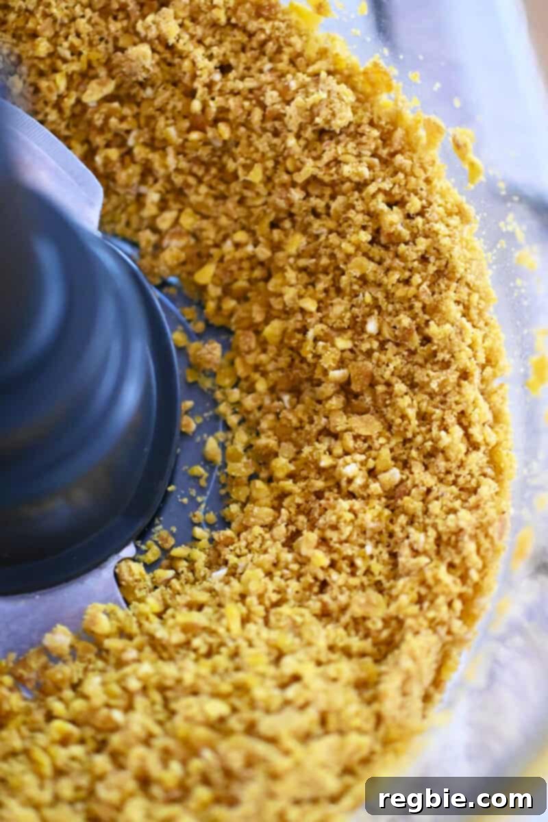 An overhead shot of an open food processor bowl containing finely crushed plantain chip crumbs, ready for breading.