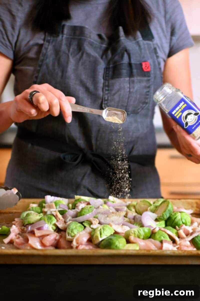 One Pan Chicken and Crispy Brussels Bake 6 A person in a gray apron carefully sprinkling a spoonful of Nomtastic Grilling Powder over the chicken, Brussels sprouts, and bacon on the sheet pan.