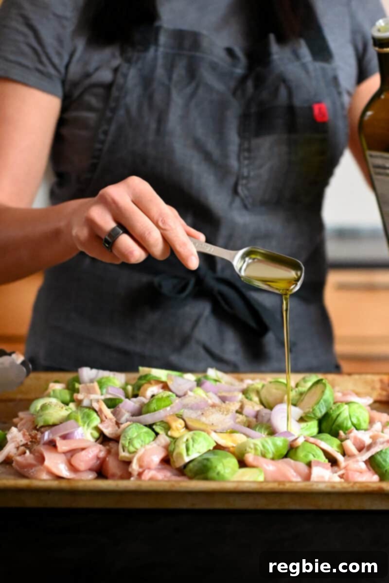 One Pan Chicken and Crispy Brussels Bake 7 A hand adding a spoonful of extra virgin olive oil to the medley of chicken and vegetables on the sheet pan before tossing.