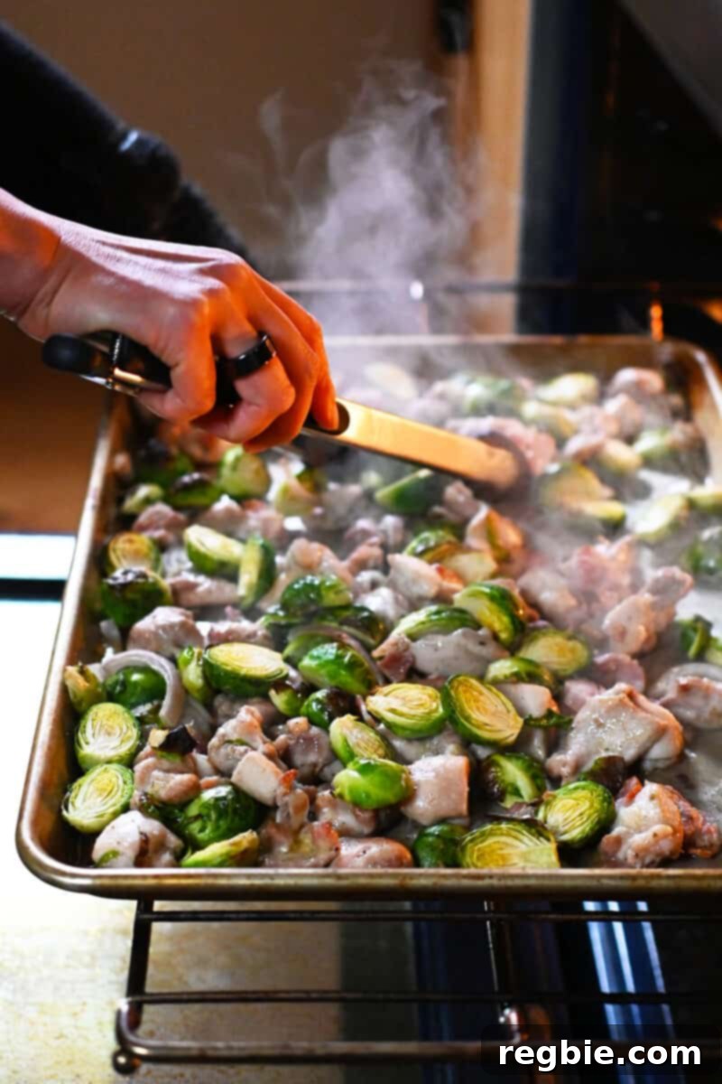 One Pan Chicken and Crispy Brussels Bake 10 Tongs tossing the ingredients on a sheet pan after 15 minutes of baking, preparing them for the final roasting stage to achieve perfect browning.