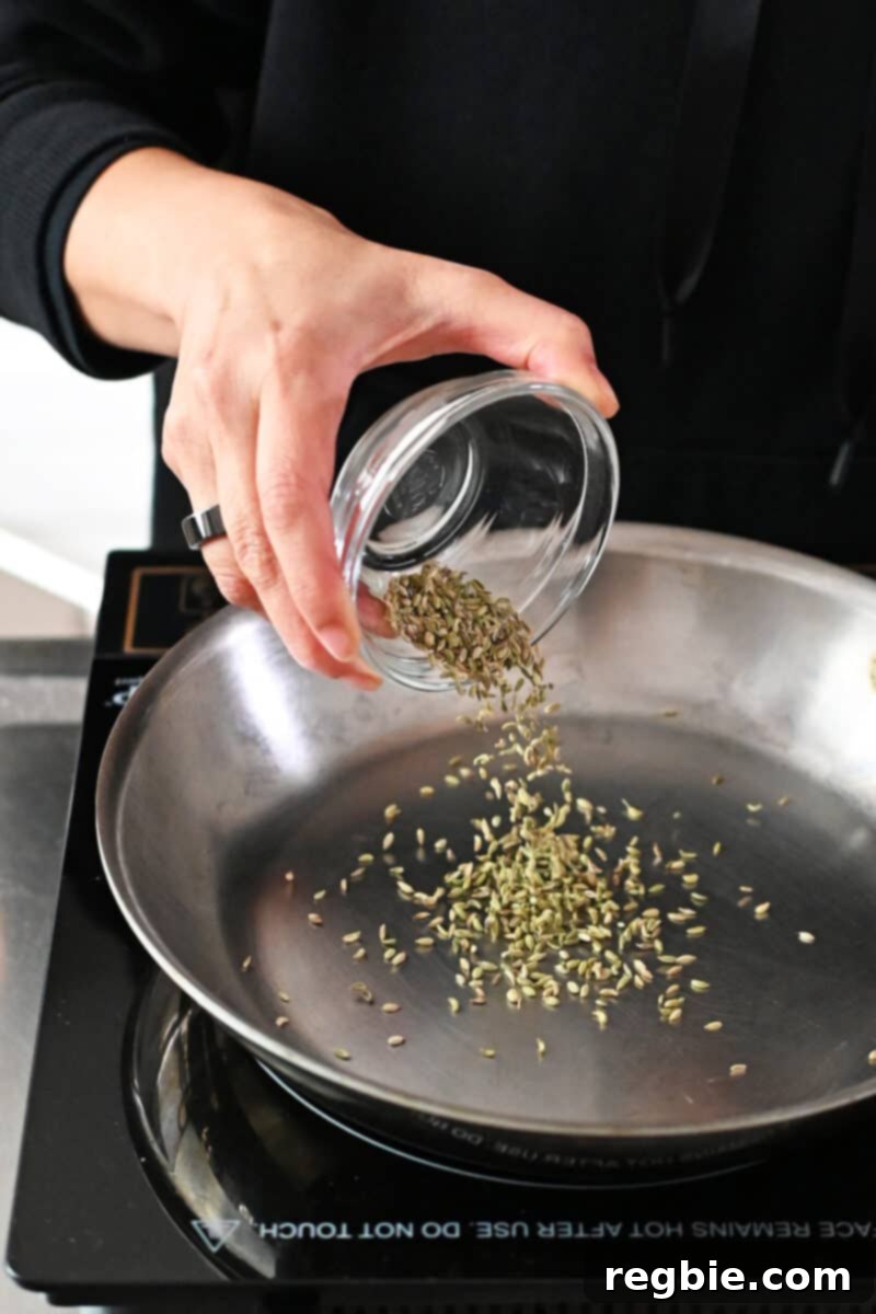 A hand is pouring fennel seeds into a stainless steel skillet.