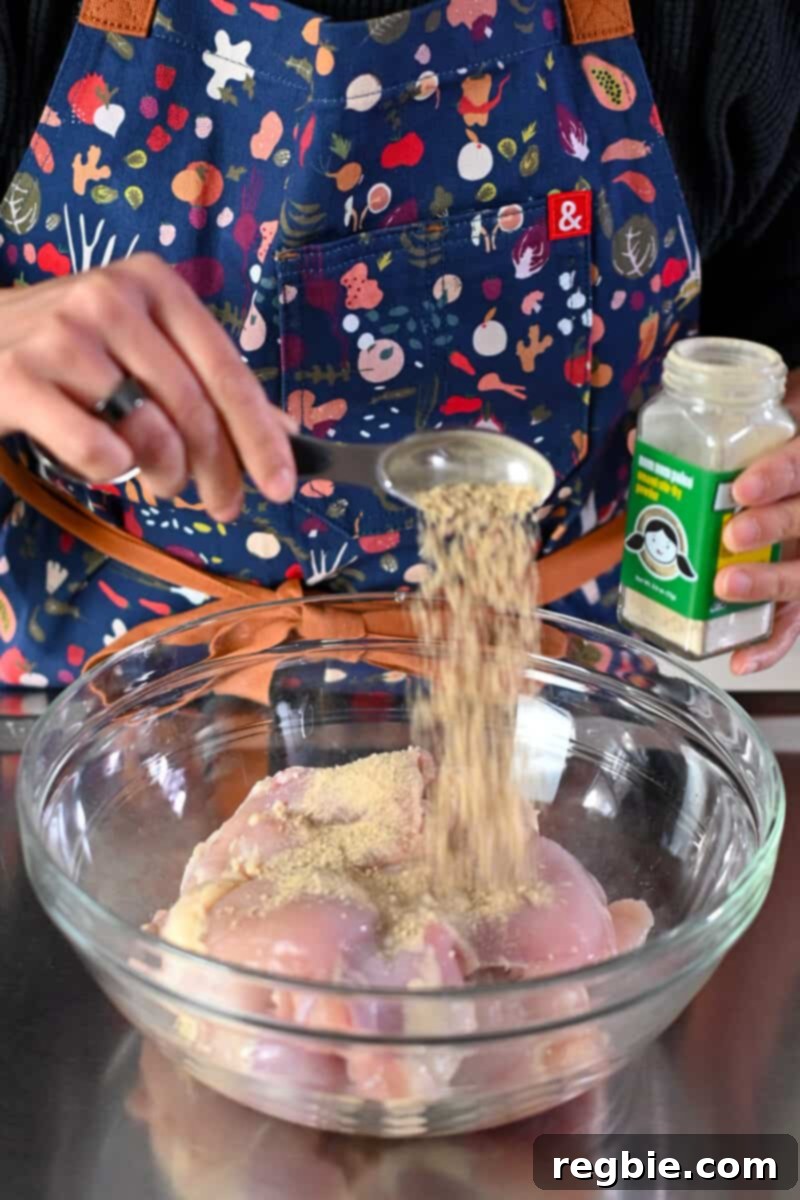 Adding a spoonful of Umami Stir-Fry Powder to a bowl of raw boneless, skinless chicken thighs for seasoning.