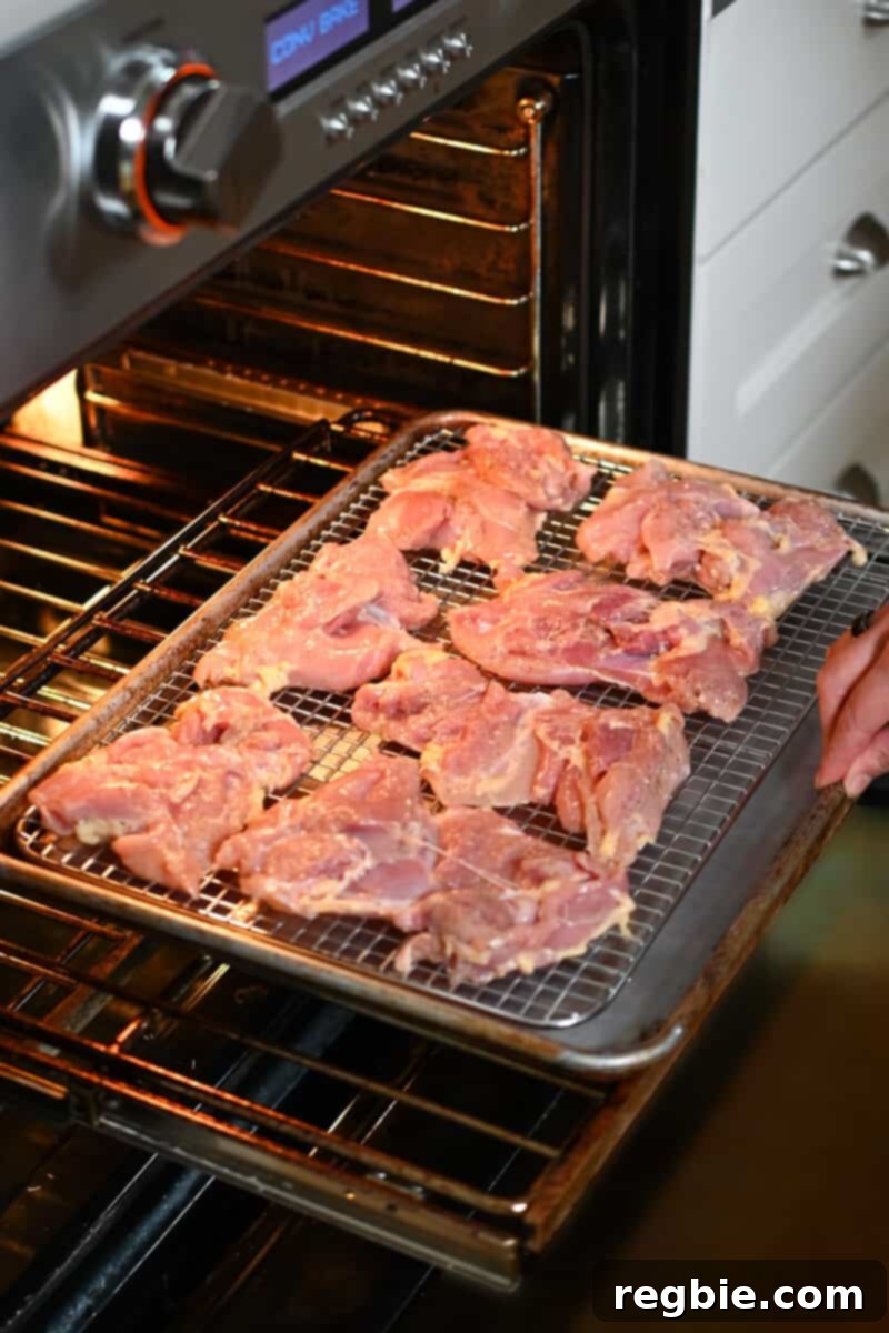 A hand is placing a baking sheet with raw Char Siu chicken into an open oven, ready for the initial baking phase.