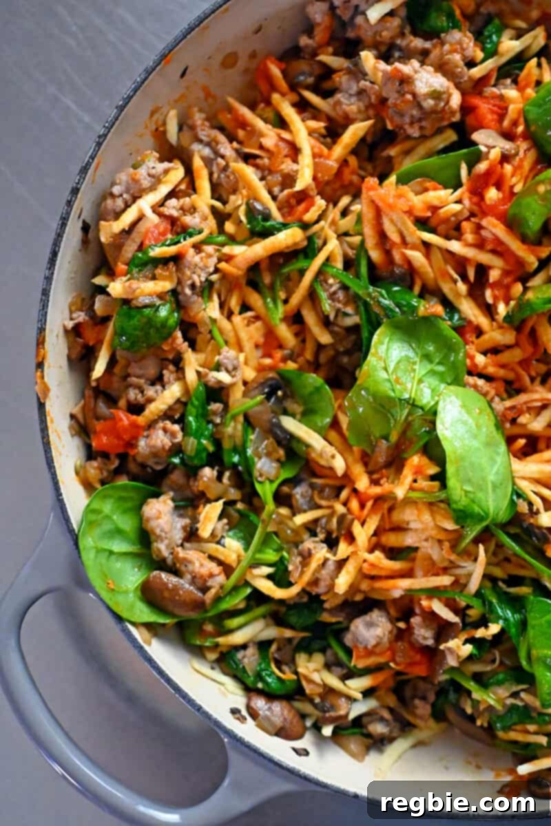 A generously filled skillet containing a perfectly combined mixture of Italian sausage, shredded white sweet potato, mushrooms, onions, and baby spinach, waiting for the egg topping.