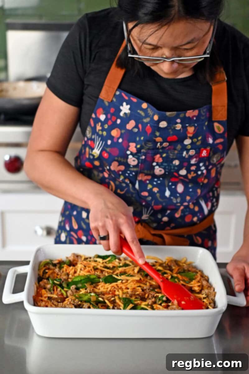 A woman in a blue apron meticulously arranging the cooked vegetables and sausage into a large baking pan, preparing for the egg mixture.