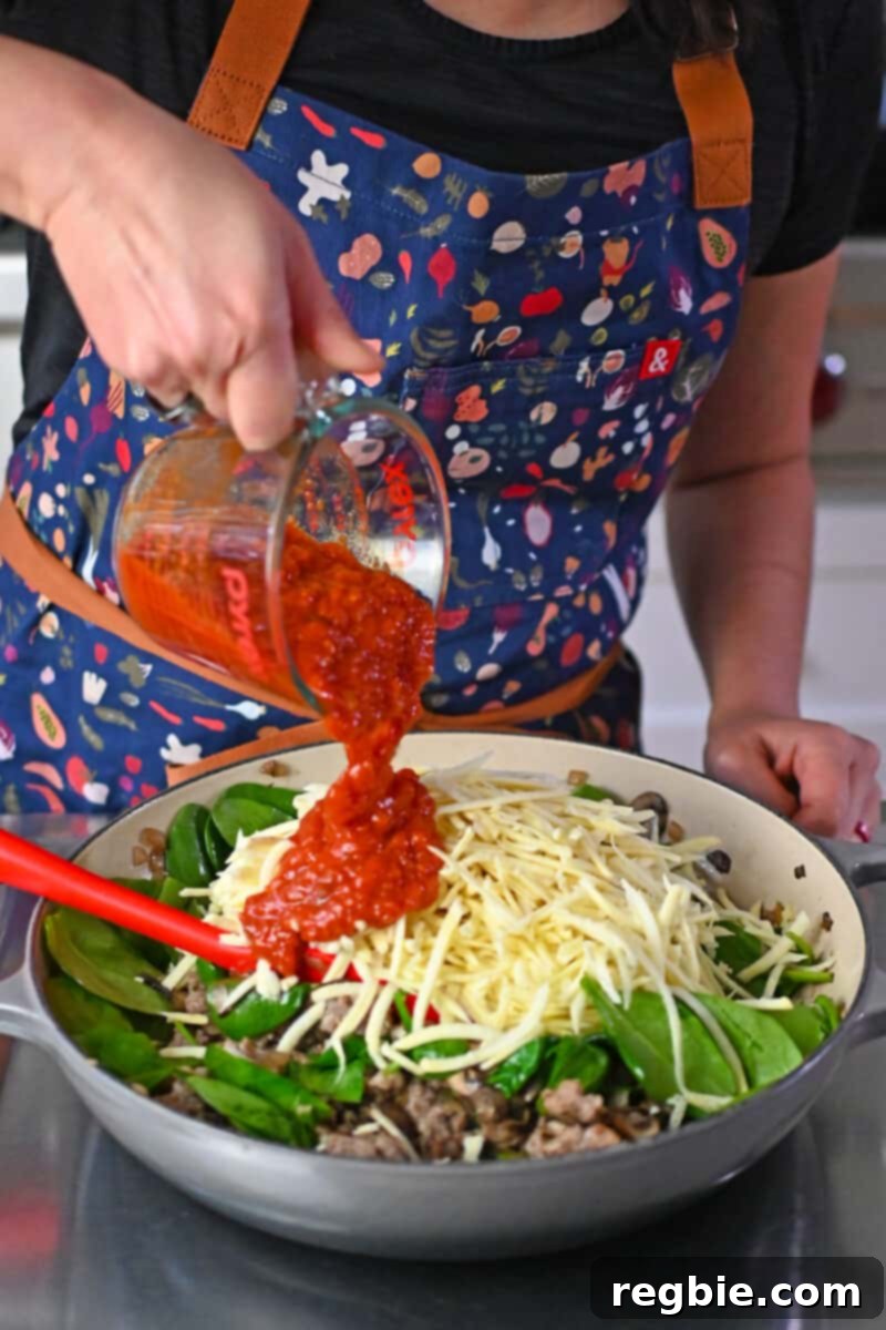 Pouring rich marinara sauce into a skillet filled with a savory blend of Italian sausage, onions, mushrooms, and shredded white sweet potatoes, ready to be mixed for the casserole base.
