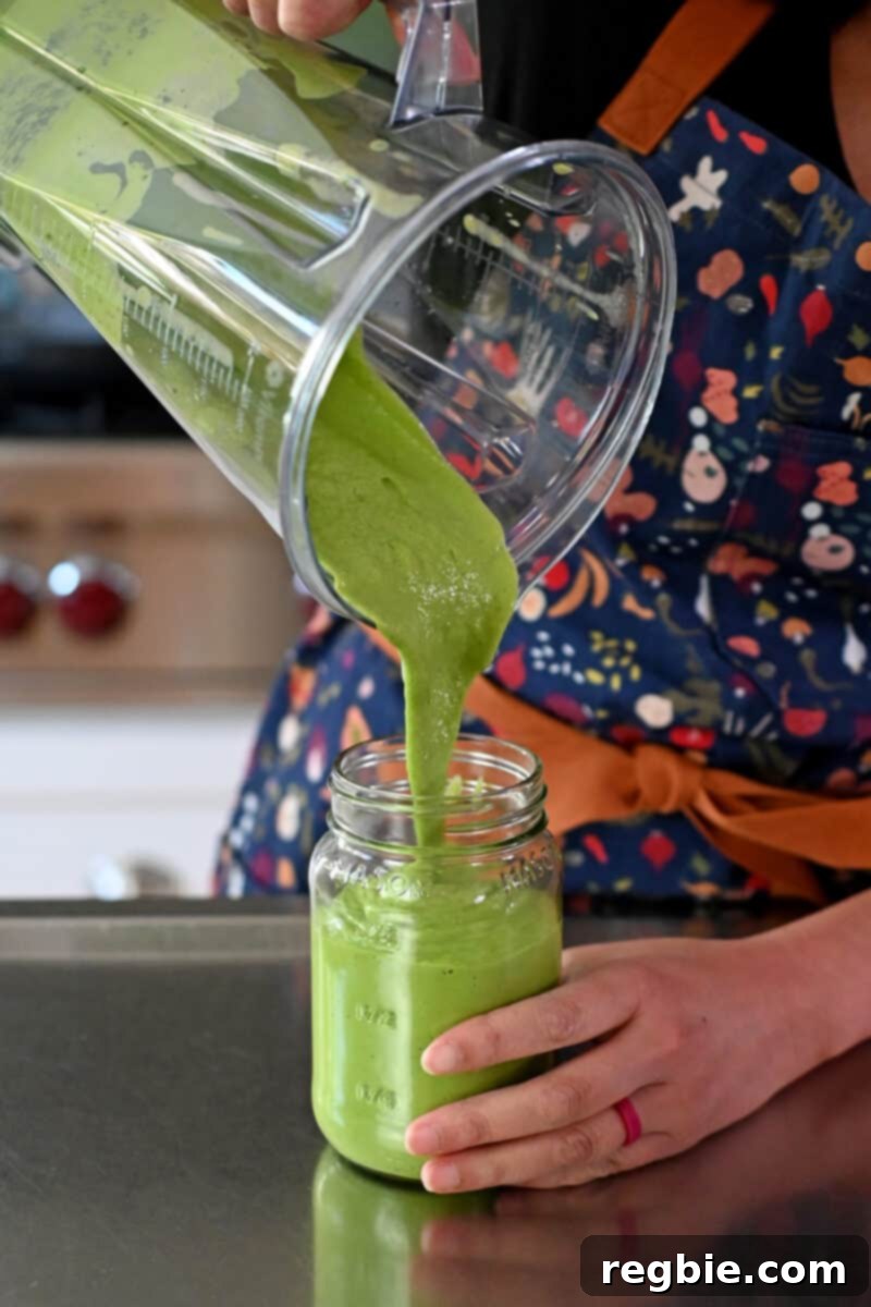 A person pouring the freshly made Green Goddess dressing into a clear glass mason jar for storage and easy serving.