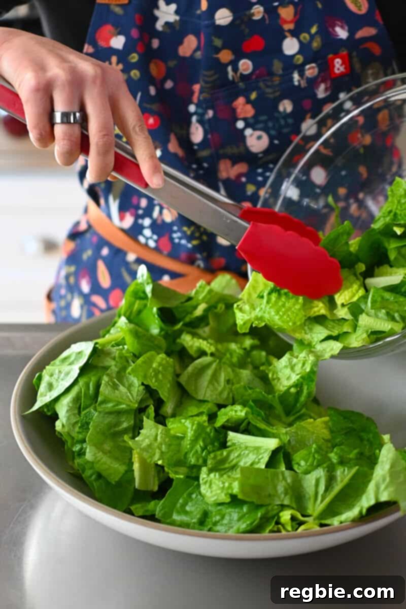 Using red tongs to carefully place fresh, chopped romaine lettuce leaves into a shallow salad serving bowl, forming the base of the Cobb salad.