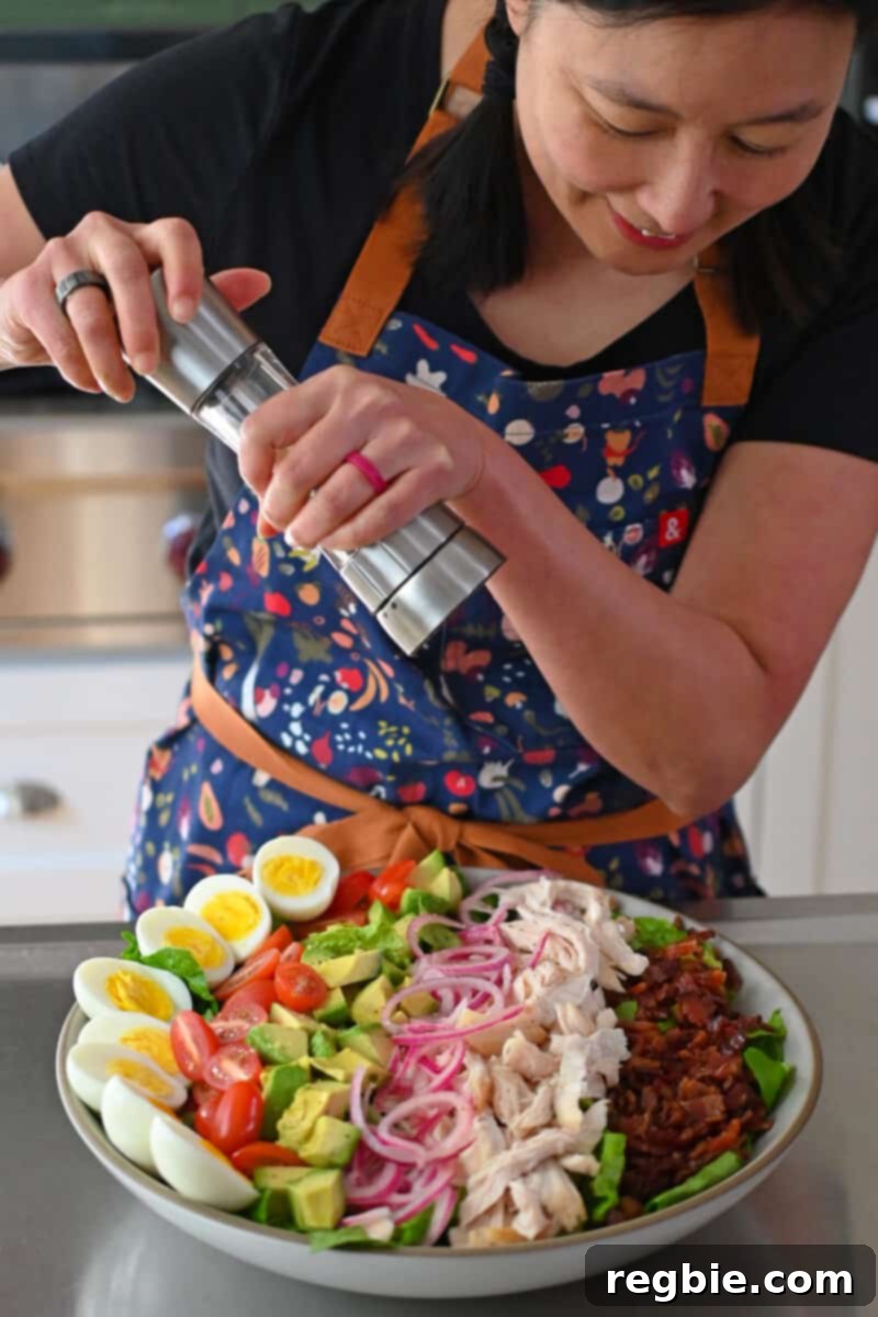An Asian woman is grinding fresh black pepper on top of a Green Goddess Cobb Salad, adding the final seasoning before dressing.