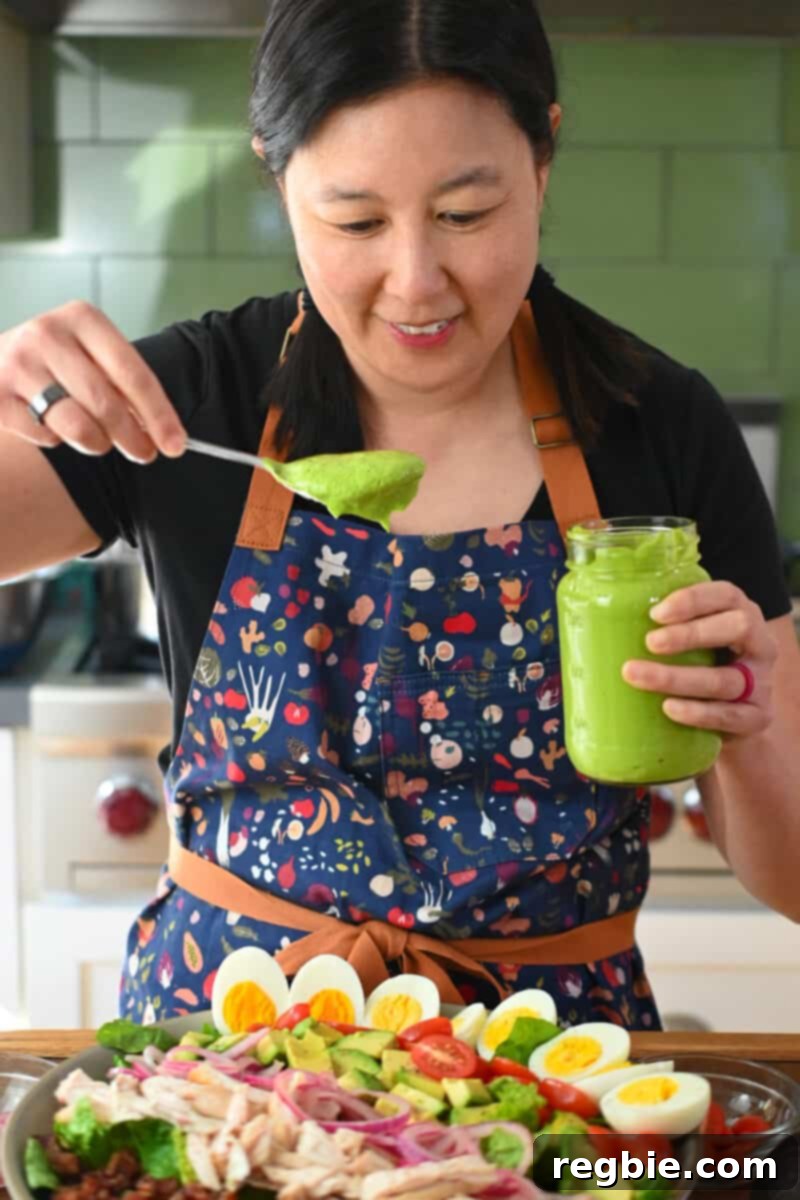 An Asian woman in a blue apron is spooning homemade Green Goddess dressing liberally over a vibrant Cobb Salad, ready to be tossed and served.