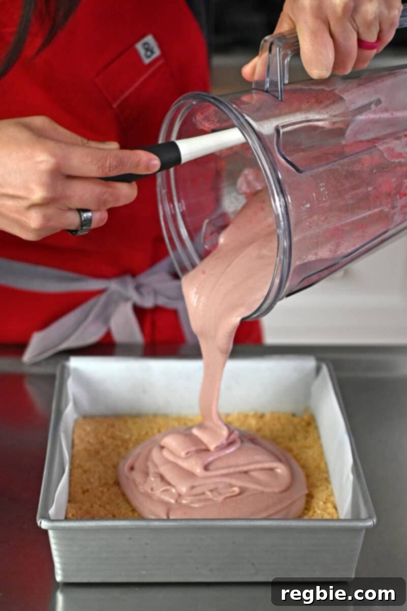 Pouring the vibrantly pink vegan strawberry cheesecake filling evenly over the pre-chilled crust in a parchment-lined baking pan.