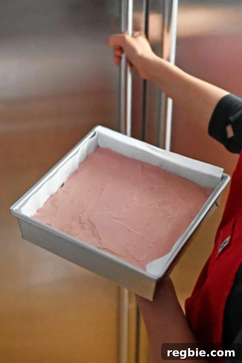 A hand gently placing a pan of freshly prepared strawberry cheesecake bars into the freezer for chilling.