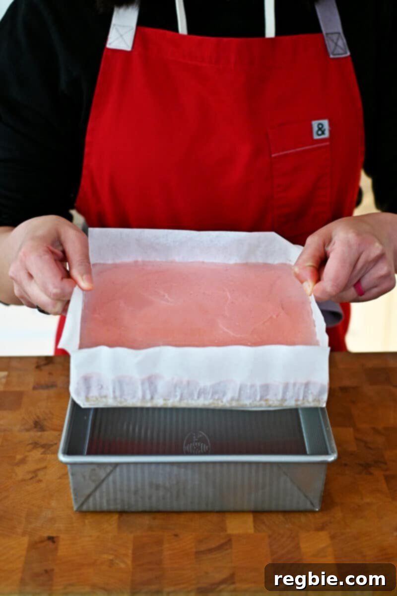 The action of carefully removing the perfectly chilled strawberry cheesecake bars from the baking pan by lifting the parchment paper overhang.