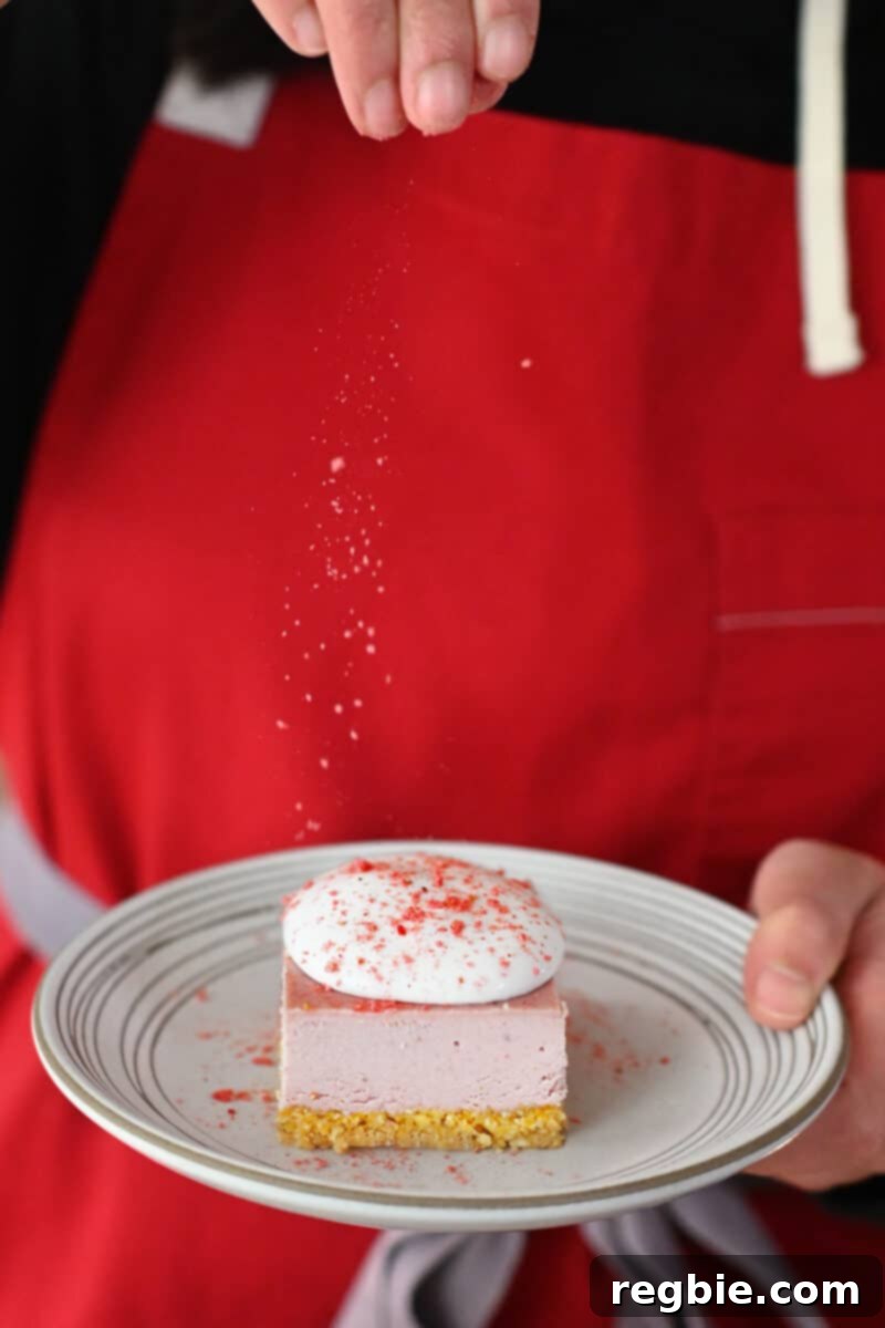 A person in a red apron presenting a white plate holding a single strawberry cheesecake bar, beautifully garnished with whipped coconut cream and a sprinkle of strawberry powder.