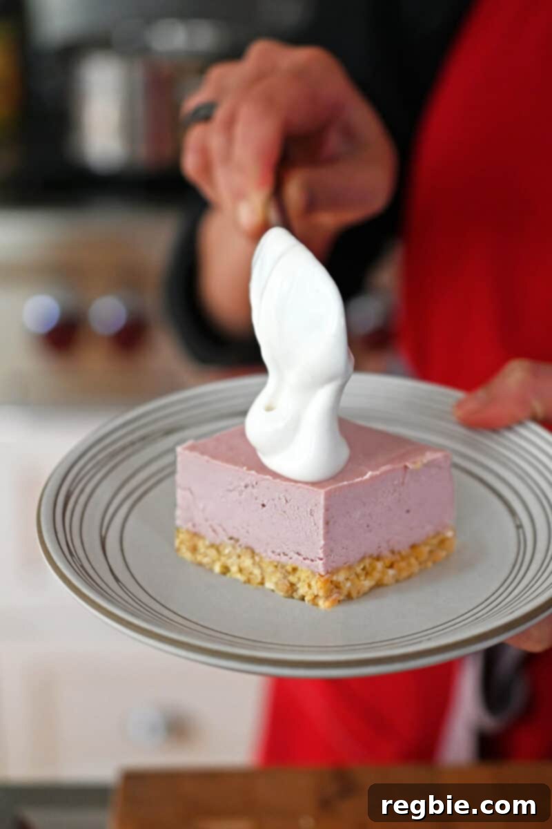 A close-up shot of a square slice of paleo and vegan strawberry cheesecake bar, being gracefully topped with a swirl of rich whipped coconut cream.