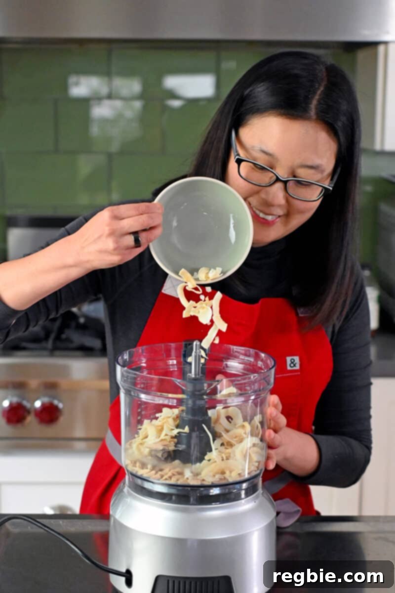 A smiling Asian woman carefully adding toasted coconut flakes into a food processor already containing toasted almond slivers, preparing for the crust.