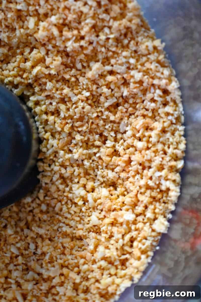 An overhead view inside an open food processor, clearly showing the toasted almonds and coconut flakes processed to a coarse cornmeal consistency, ideal for the crust.