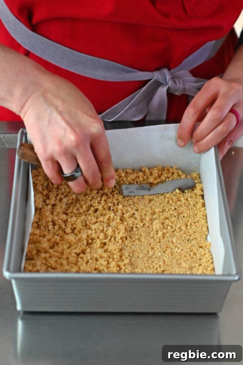 A hand expertly using an offset spatula to firmly press the coconut and almond crust into an even layer on the bottom of a parchment-lined square baking sheet.