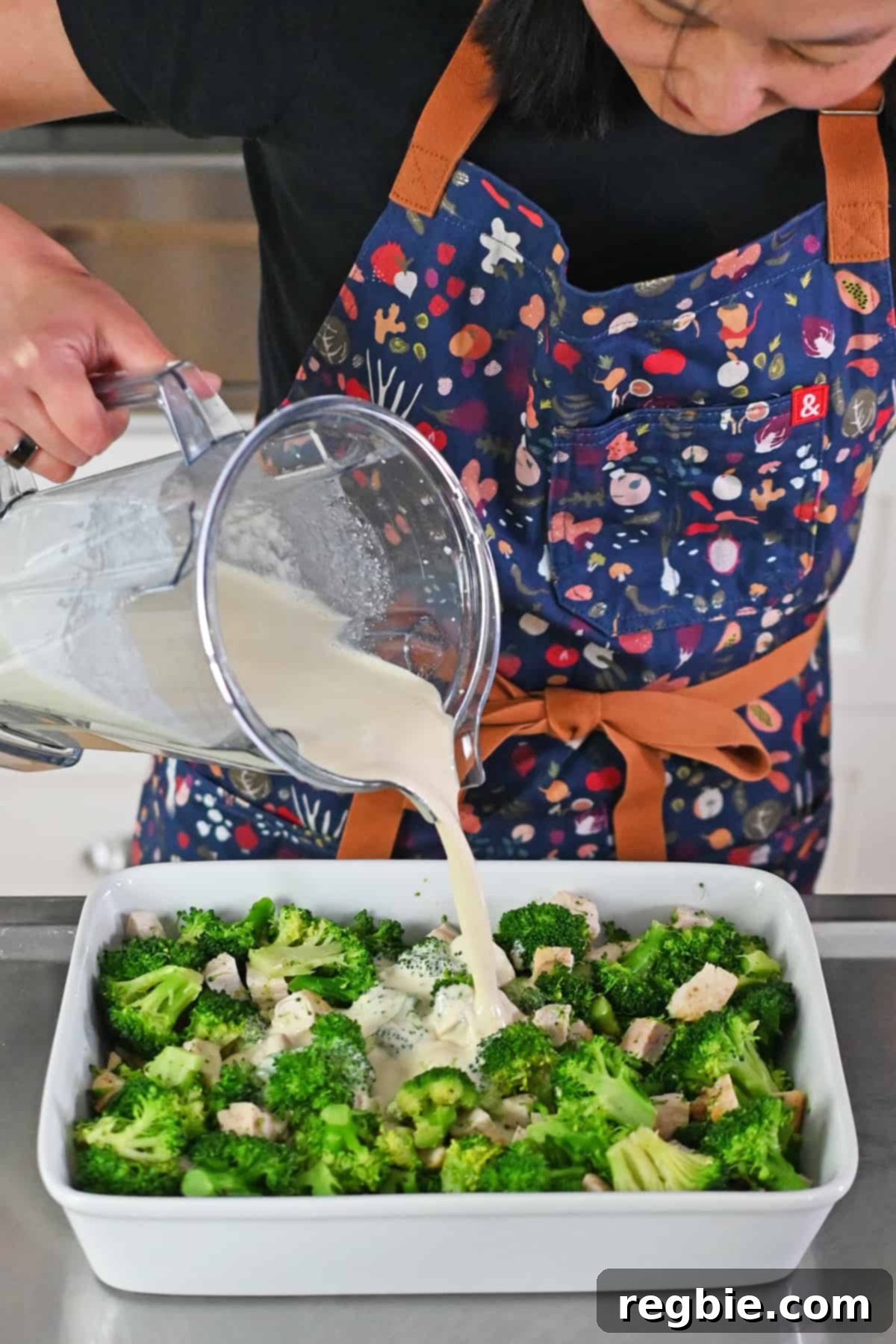 An Asian woman in a blue apron is pouring a white, creamy sauce from a blender onto the prepared broccoli and chicken in a white casserole pan.