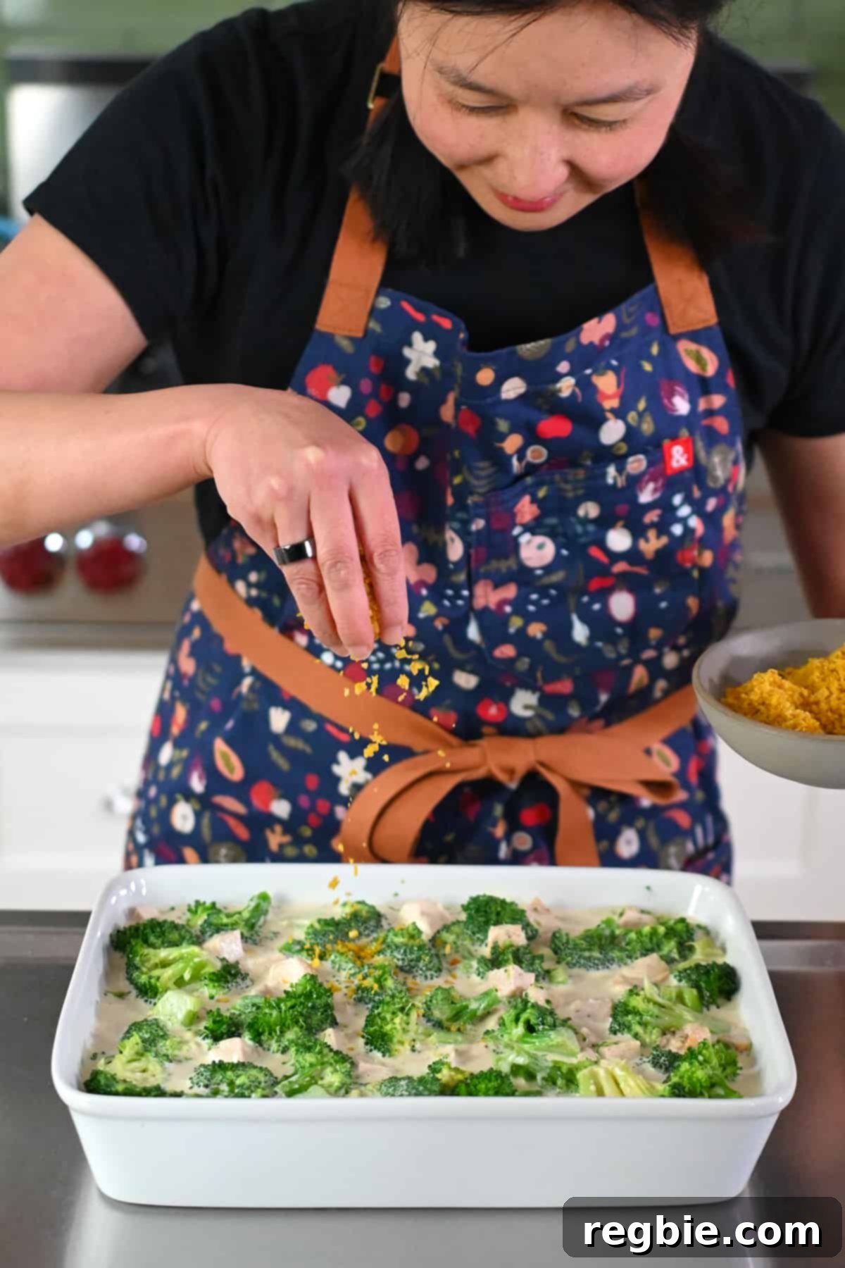 An Asian woman is sprinkling crushed plantain chips onto an unbaked paleo chicken divan casserole, adding a delightful crunchy topping.