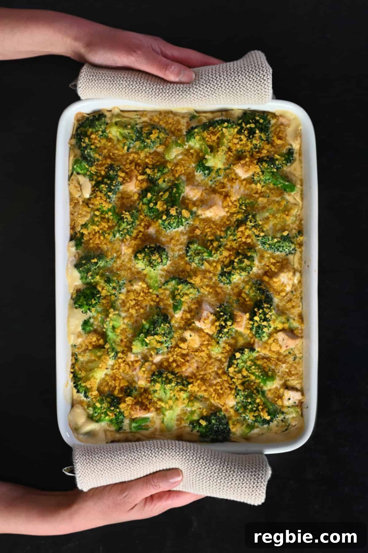 Two hands are holding a freshly baked paleo chicken divan casserole right out of the oven, steam gently rising.