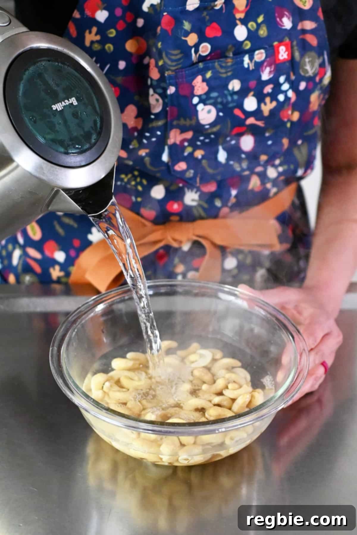 Pouring boiling water from an electric kettle into a clear glass bowl with raw cashews for soaking.