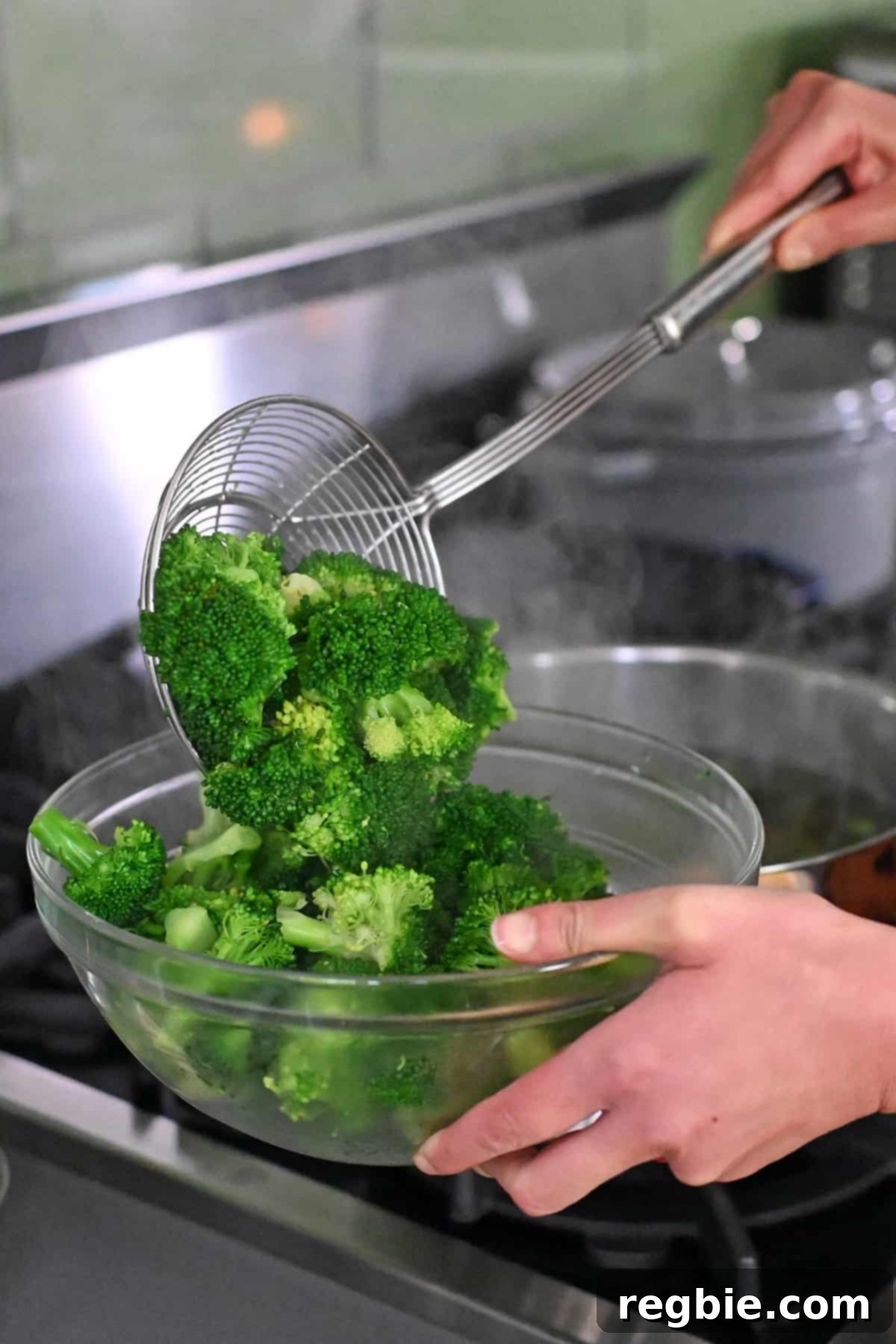 A metal sieve is being used to transfer cooked broccoli from a pot of boiling water to a clear glass bowl, preserving its vibrant green color.