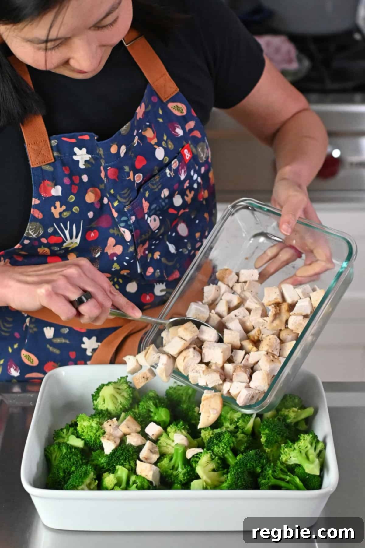 An Asian woman in a blue apron is adding cooked cubed chicken to a casserole dish with bright green cooked broccoli florets.