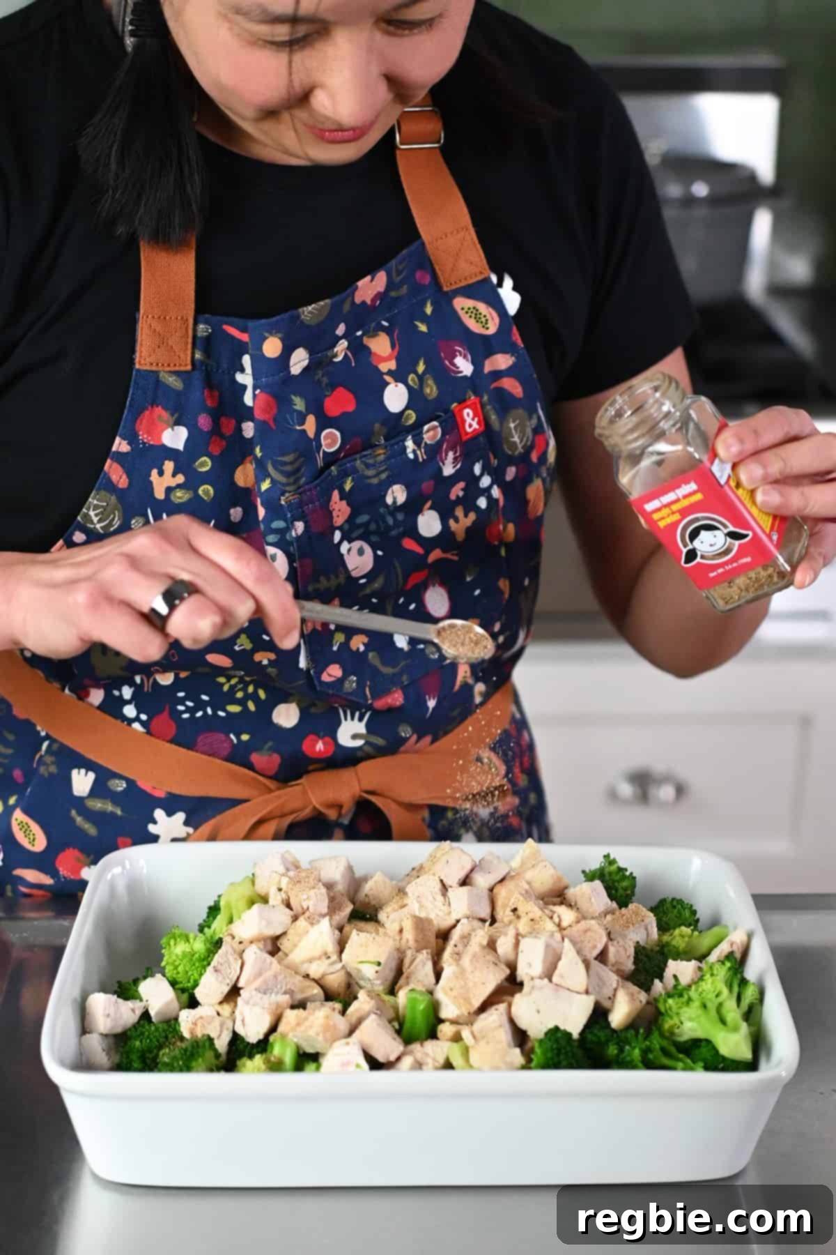 An Asian woman is adding some Nom Nom Paleo's Magic Mushroom Powder to a casserole dish containing cooked chicken cubes and broccoli.
