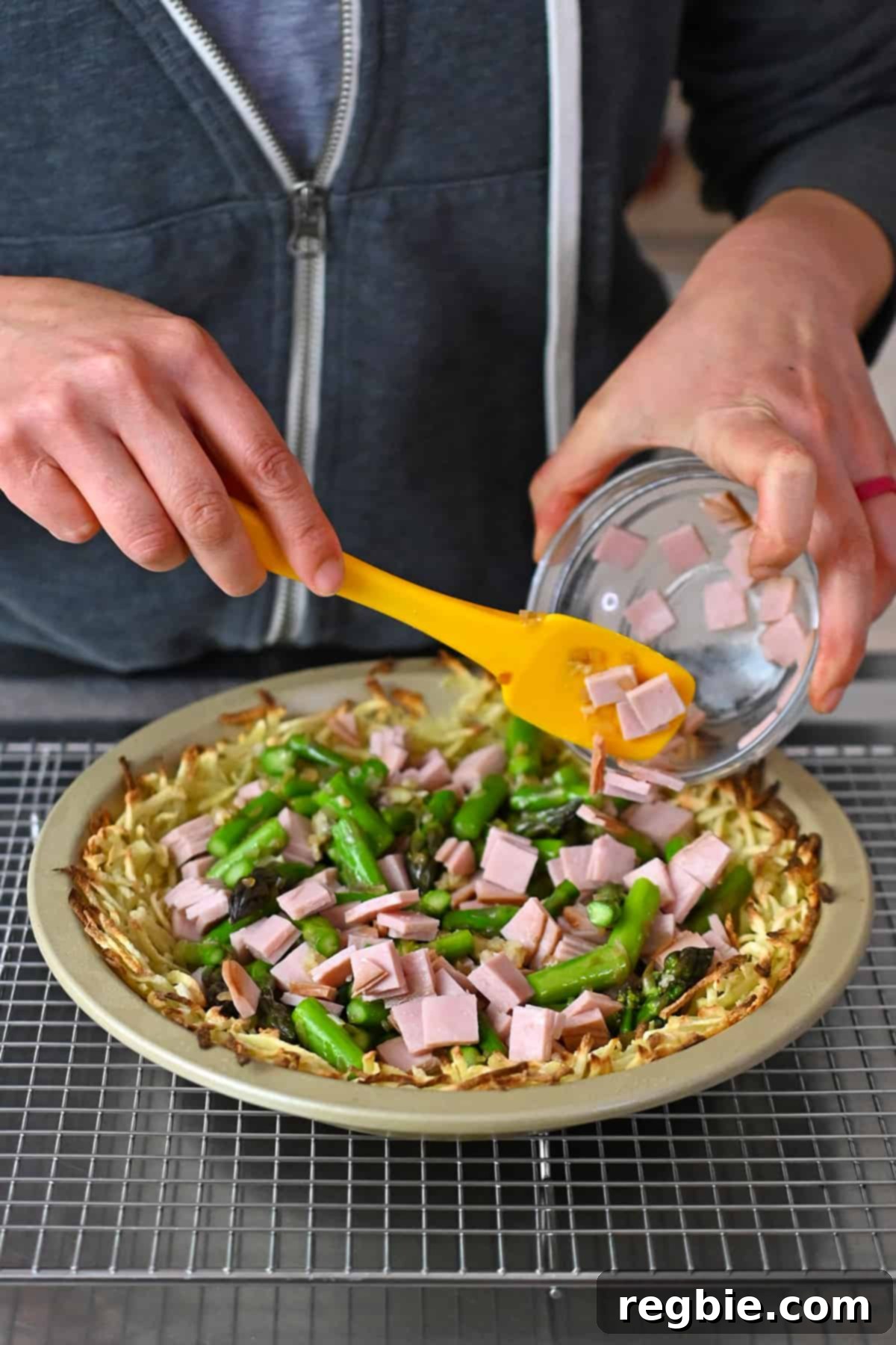 The assembled quiche filling with cooked asparagus and diced ham being added to the baked shredded potato crust in a pie plate.